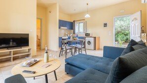 Una sala de estar con televisor, sofá y mesa de centro. Se observa una cocina al fondo con una mesa de comedor y sillas.