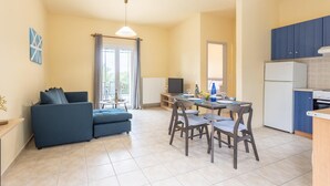 Un espacio habitable compacto con sofá, mesa de comedor, sillas, área de cocina con armarios y electrodomésticos, y un ventanal con cortinas.
