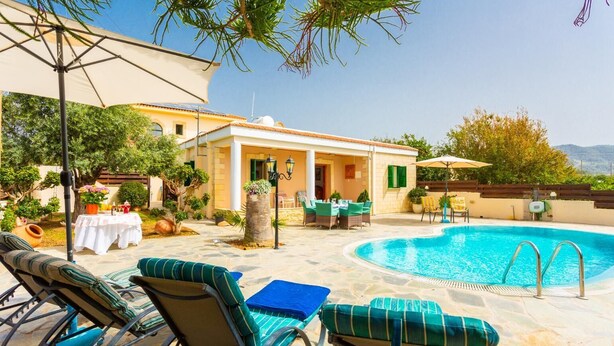A poolside area with lounge chairs, a table, and a small building with a door. - Villa Constantinos - Three Bedroom Villa, Sleeps 6 (Agia Marina Chrysochous)