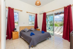 Un dormitorio con cama, dos mesitas de noche, un ventanal y un balcón con cortinas rojas.