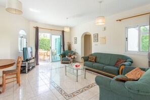 Amplia sala de estar con sofá verde, mesa de centro de vidrio y televisor sobre un mueble. Hay una mesa de comedor con sillas y vista al exterior a través de una puerta de vidrio.