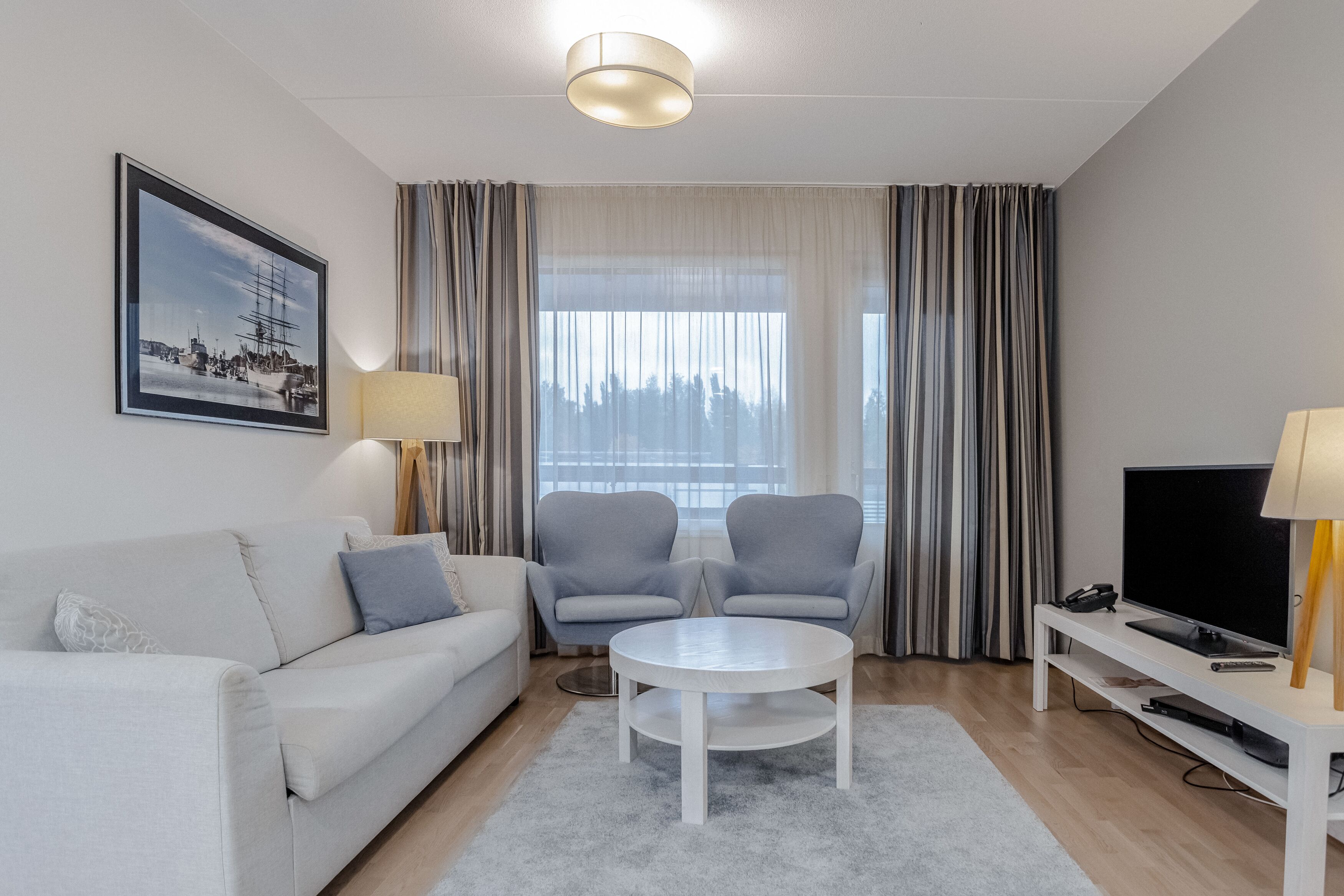 Uma sala de estar moderna com um sofá branco, duas poltronas cinzas, uma mesa de centro redonda, uma TV sobre um suporte branco e uma grande janela com cortinas.