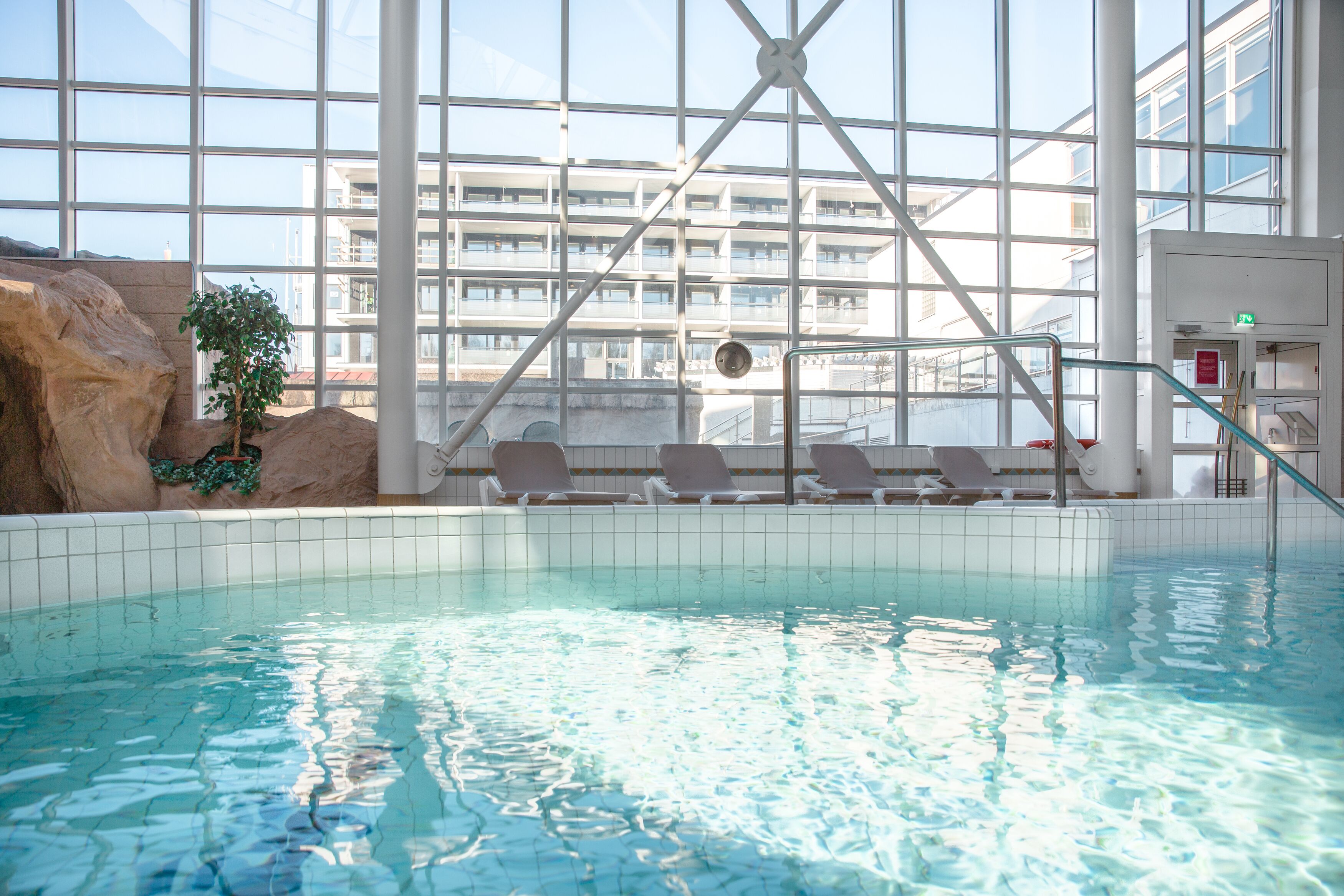 Una piscina interior con agua cristalina, iluminada por grandes ventanales y vistas a un edificio moderno.
