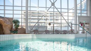 Una piscina interior con agua cristalina, iluminada por grandes ventanales y vistas a un edificio moderno.
