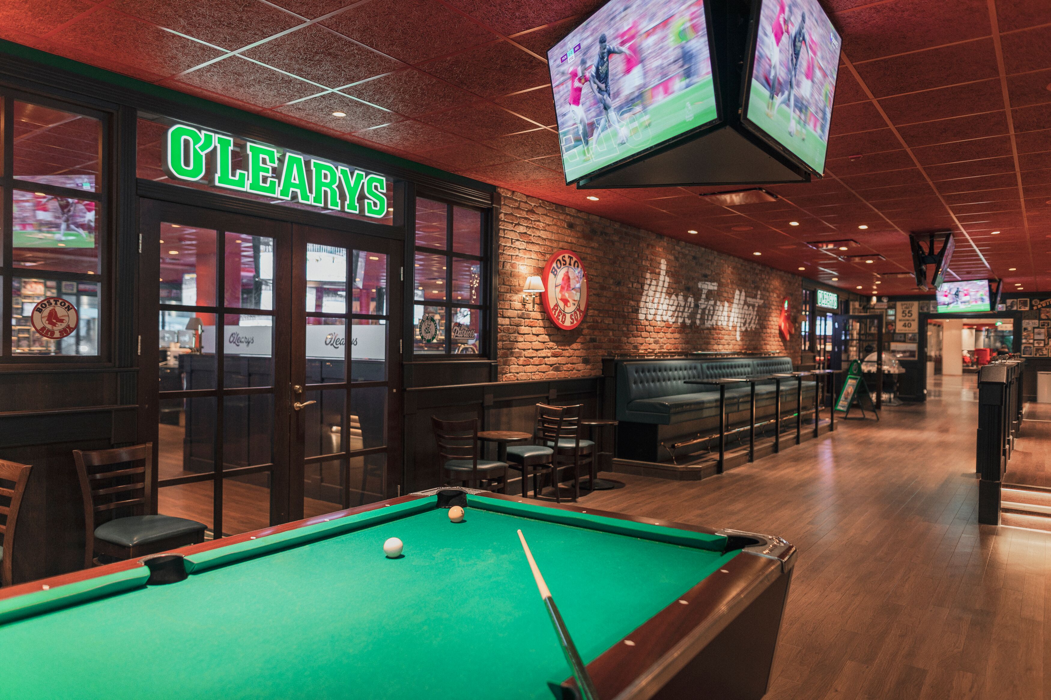 A billiards table with a pool cue and balls, a brick wall with a Boston Red Sox sign, and a bar area with seating.