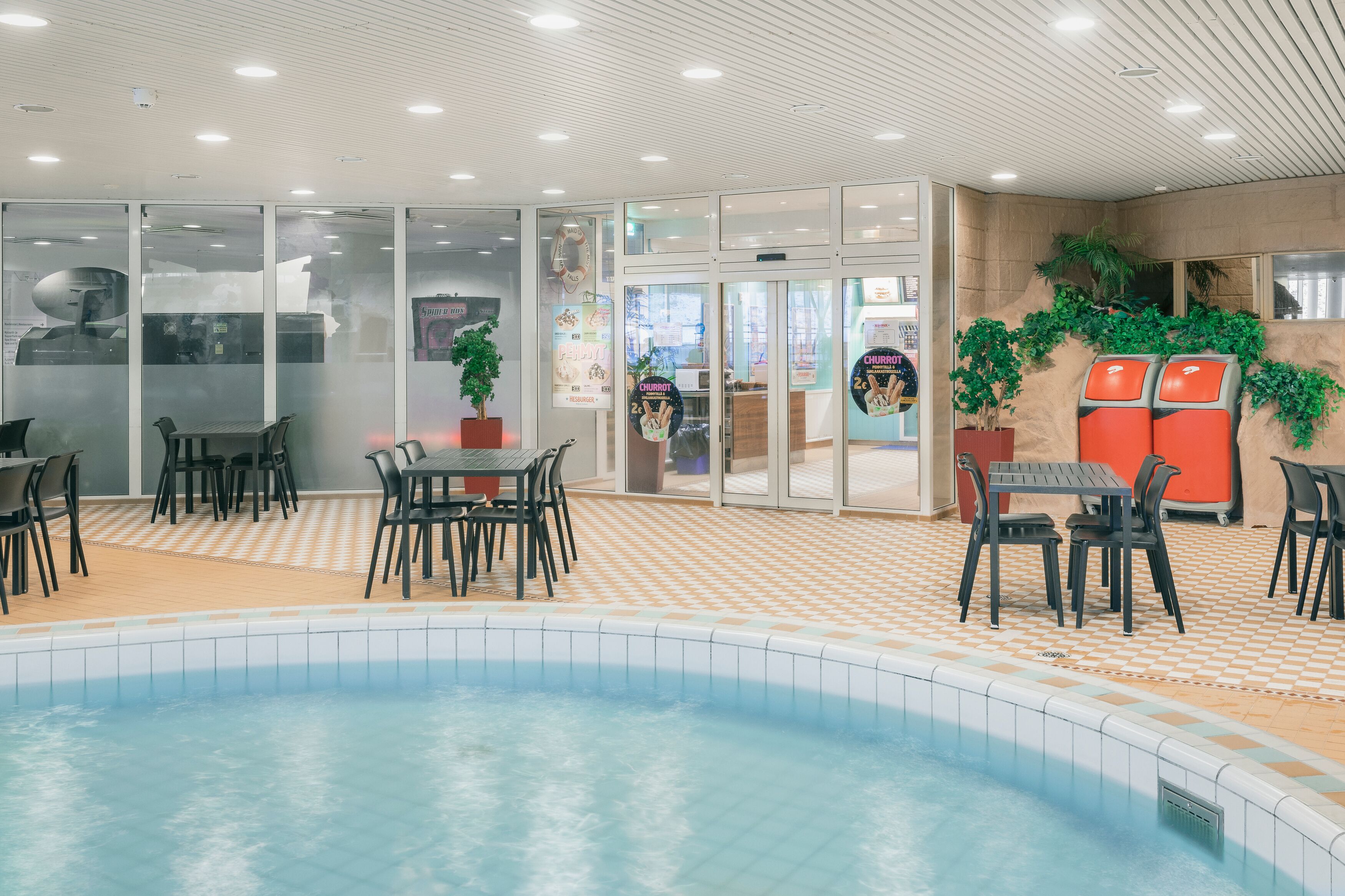 Un área de piscina interior con suelo de baldosas, zonas de estar y puertas de cristal que dan a un restaurante.