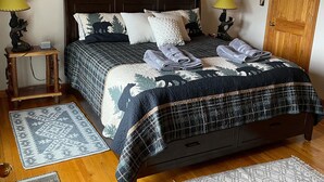 A bedroom with a wooden ceiling, a bed with a dark quilt, two bedside tables with lamps, a wooden door, and a wall-mounted painting. - Paradise Peak - Tranquility and majestic splendor! (Butler)