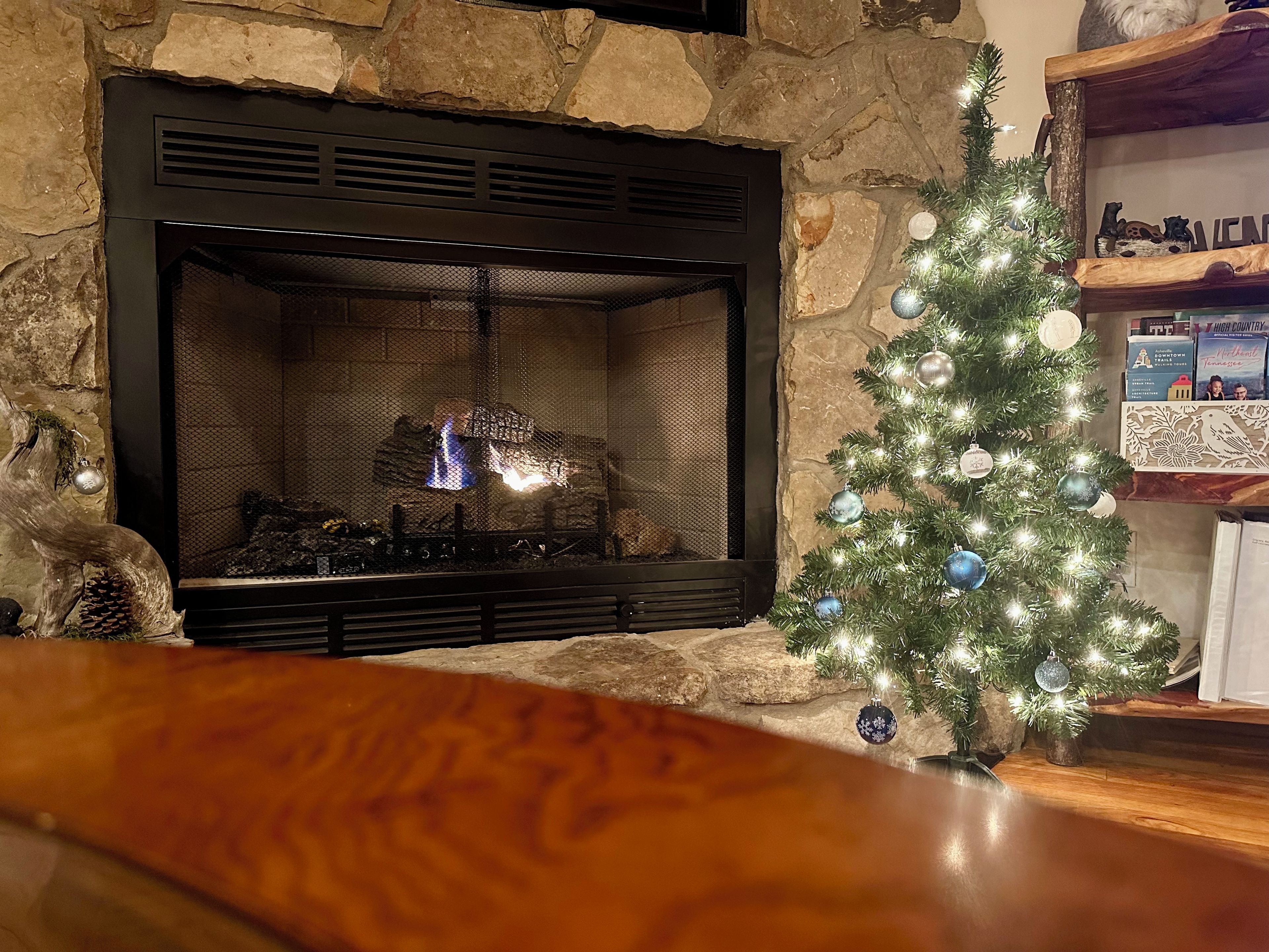 A fireplace with a stone wall backdrop, a lit fire, and a decorated Christmas tree.