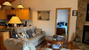A cozy living room with a sofa, a coffee table, and a fireplace. There is a kitchen area with wooden cabinets and a lamp. - Paradise Peak - Tranquility and majestic splendor! (Butler)