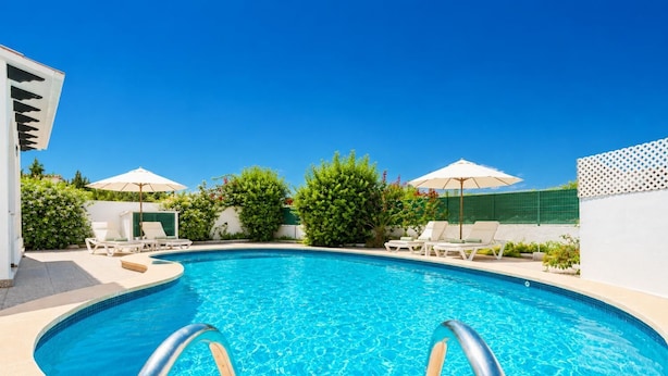 A swimming pool with lounge chairs and umbrellas, surrounded by greenery and a clear sky.