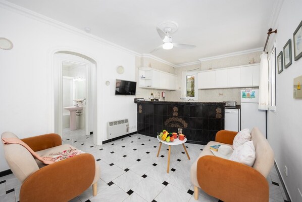 Uma sala de estar moderna com dois sofás, uma mesa de jantar com frutas, uma televisão e uma cozinha com armários brancos e painel de azulejos pretos.