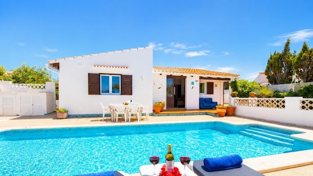 A swimming pool with lounge chairs and a table set for two, a small building with a door and windows, and a clear blue sky. - Villa Ca Nostra - Three Bedroom Villa, Sleeps 6 (Cala en Porter)
