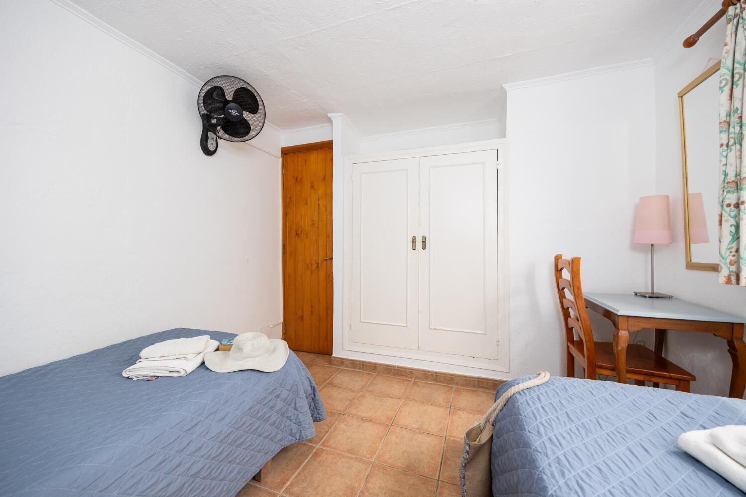 Une chambre avec deux lits, un bureau, une chaise, un ventilateur et une armoire.