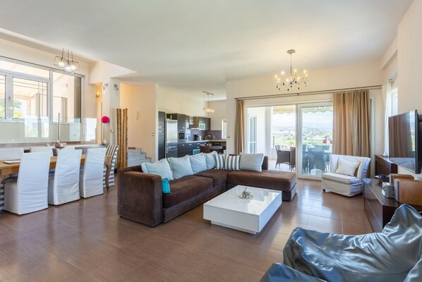 Ein großzügiger Wohnbereich mit einem großen, braunen Ecksofa, einem weißen Couchtisch und einem Essbereich mit gestreiften Stühlen.