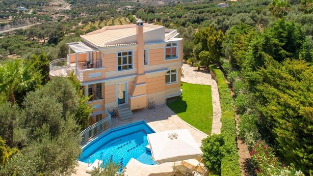 Une maison à plusieurs étages avec une piscine, entourée d’une végétation luxuriante et de collines.