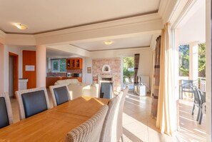 A spacious living area with a wooden dining table, comfortable seating, a kitchen in the background, and a balcony with outdoor furniture. - Villa Pelagos - Five Bedroom Villa, Sleeps 10 (Agia Triada)