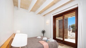 Ein Schlafzimmer mit Bett, Nachttischlampe, Ventilator und Blick auf einen Außenbereich mit Pool und Sonnenschirmen.