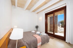 A bedroom with a bed, a bedside lamp, a fan, and a view of an outdoor area with a pool and umbrellas. - Water Front Villa Shalom (Binibeca)
