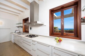 Uma cozinha moderna com armários brancos, fogão a gás, coifa de aço inoxidável e uma janela grande com vista para o mar.