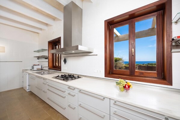 Una cocina moderna con muebles blancos, estufa a gas, campana de acero inoxidable y un amplio ventanal con vista al mar.