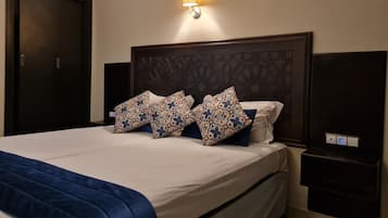 Habitación de hotel con una cama grande, cabecera oscura, almohadas estampadas, una lámpara de noche y una mesita.