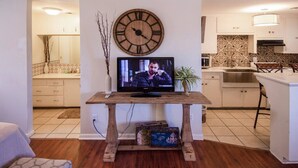Un salon avec un meuble TV en bois, une grande horloge murale et une cuisine en arrière-plan.