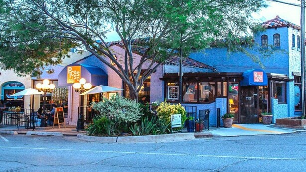 Une vue d’une rue où l’on aperçoit un restaurant avec une terrasse, un bâtiment bleu doté d’un toit en tuiles et un arbre au premier plan.