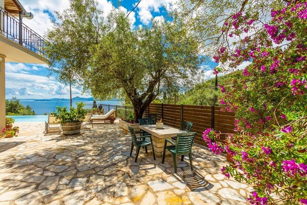 A stone patio with a table and chairs, overlooking a body of water and mountains. - Villa Katerina - Five Bedroom Villa, Sleeps 10 (Nisaki)