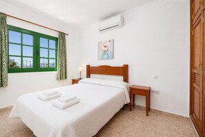 A hotel room with a bed, a nightstand, an air conditioning unit, a window with curtains, and a painting on the wall. - Villa Alegranza - Three Bedroom Villa, Sleeps 6 (Puerto del Carmen)