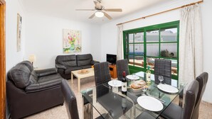 Un soggiorno moderno con un tavolo da pranzo in vetro, divani in pelle, una ventola a soffitto e una vista attraverso una finestra con cornice verde.