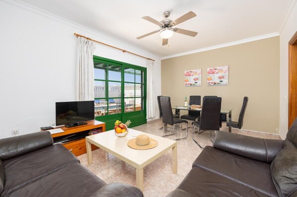 A living room with a TV, a dining table with chairs, a ceiling fan, and a view of a pool through a green door.