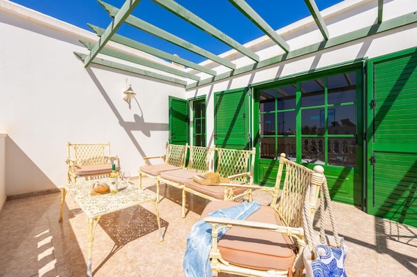 Ein sonniger Außenbereich mit Korbmöbeln und einem Tisch, grünen Rollläden und einer Pergola.
