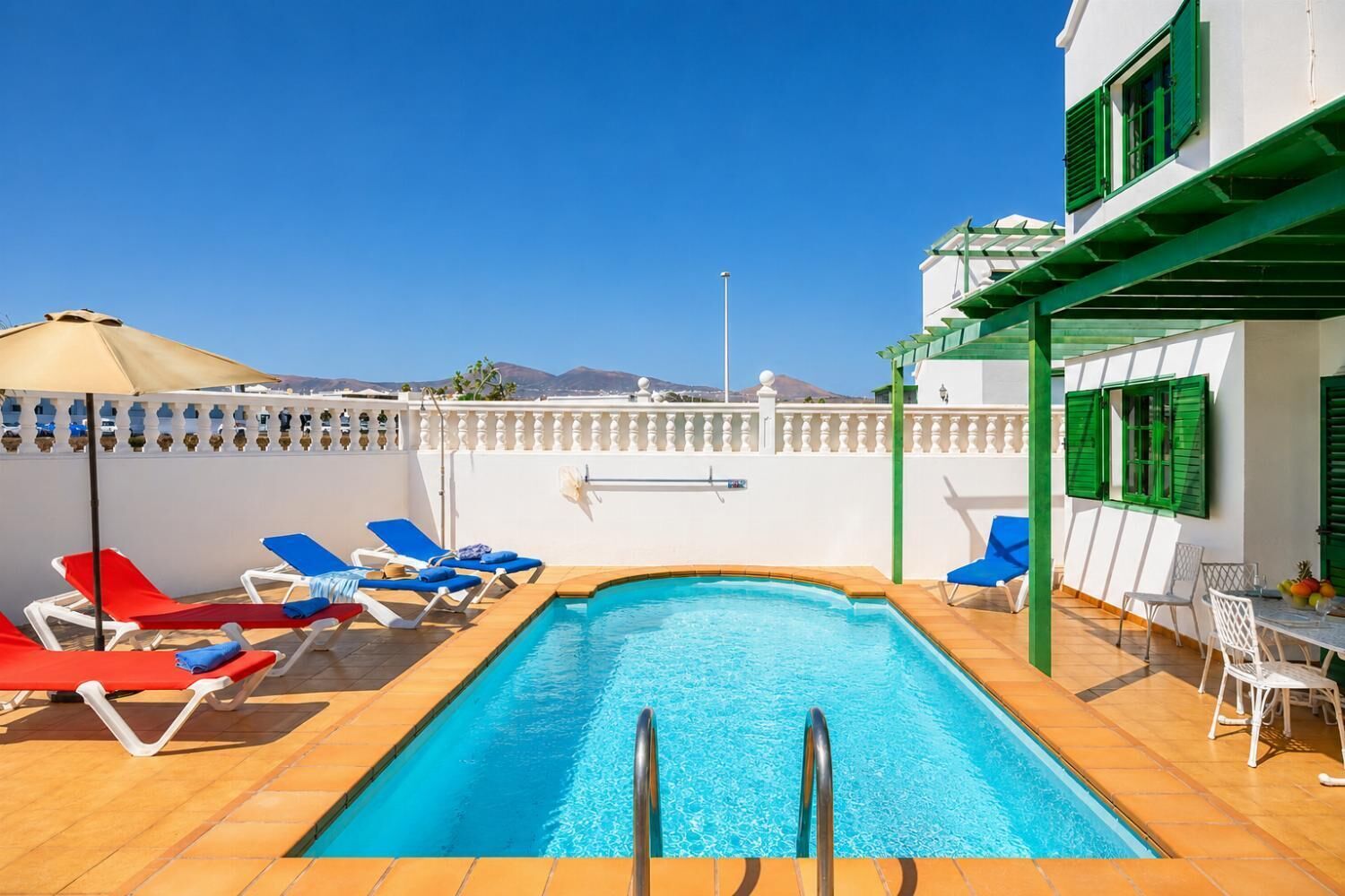 A swimming pool with sun loungers and a table with chairs, surrounded by a white wall with green shutters.