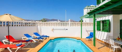 A swimming pool with sun loungers and a table with chairs, surrounded by a white wall with green shutters.
