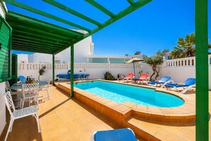 Piscina com pergolado, espreguiçadeiras e área de refeições com cadeiras e mesa.