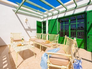 Un espacio al aire libre con mobiliario de mimbre, una mesa, contraventanas verdes y una pérgola.