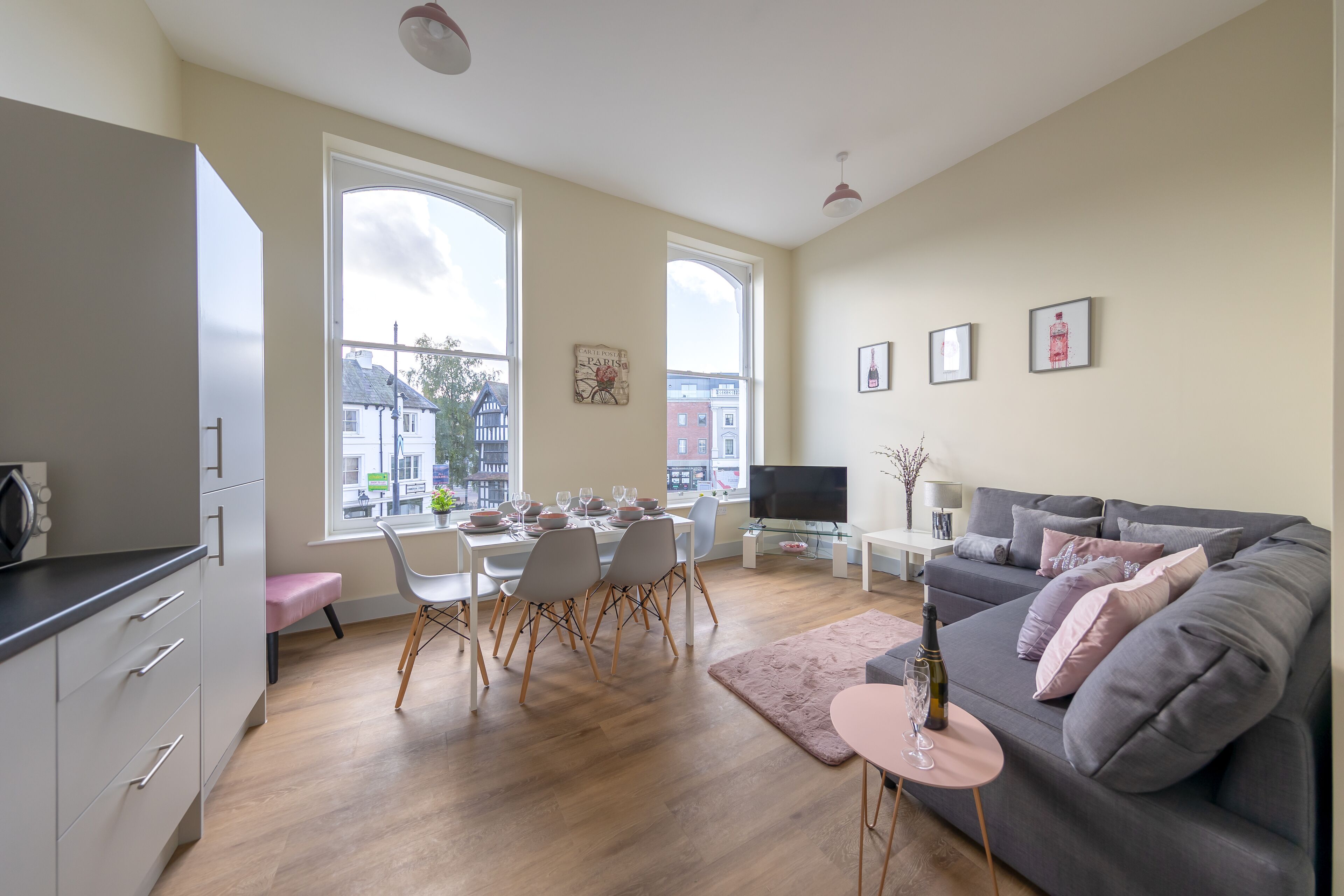 Un salon moderne avec un canapé gris, un coin repas avec une table dressée pour quatre personnes et une cuisine avec des meubles gris.
