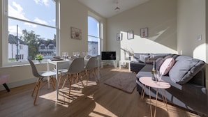 Ein geräumiges Wohnzimmer mit großem Fenster, einem grauen Sofa, einem Essbereich mit Stühlen und einem Fernseher.