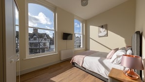 Ein Schlafzimmer mit großen Fenstern, einem Bett, einem Nachttisch mit Lampe, einem an der Wand montierten Fernseher und einer Heizung.