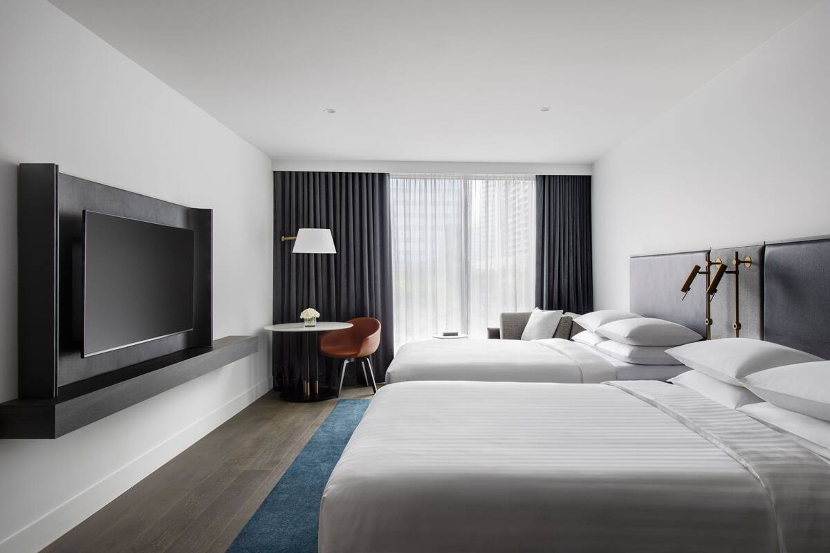 A modern hotel room with two beds, a flat-screen TV mounted on the wall, a desk with a chair, and a large window with curtains.