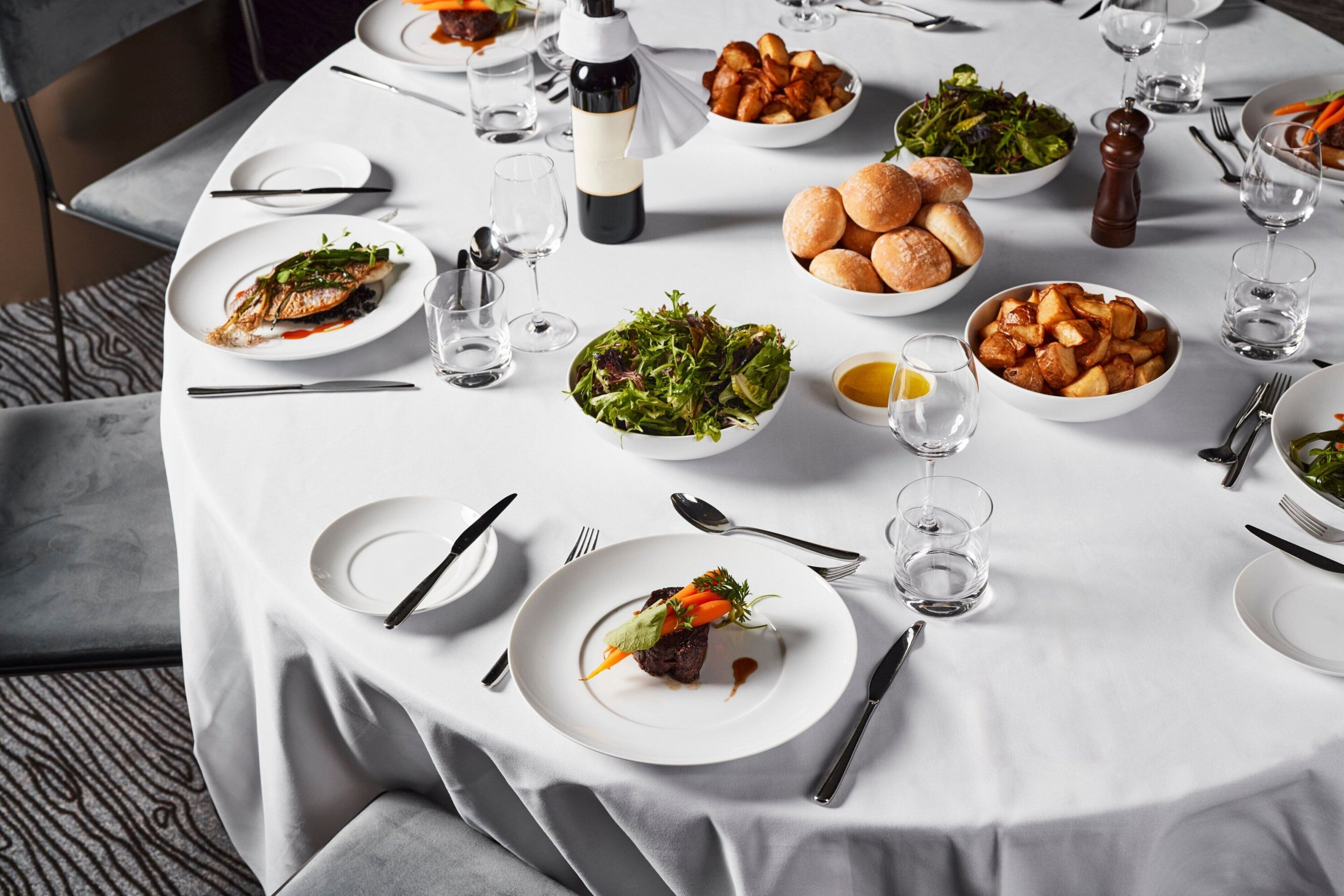 A round dining table set with various dishes, including a fish, salad, and roasted potatoes, along with wine glasses and cutlery.