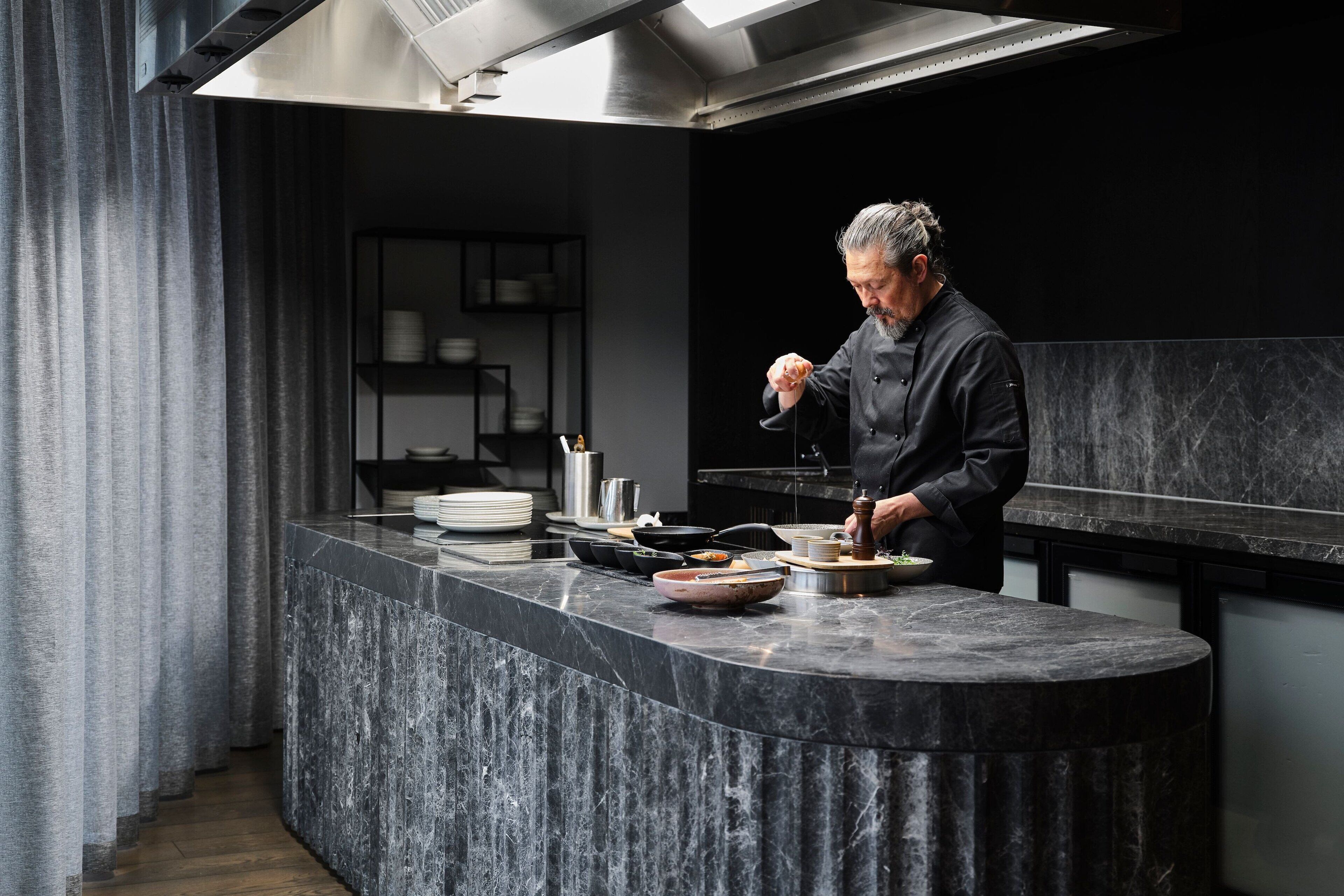 A modern kitchen with a marble countertop, a chef preparing food, and a dark interior.
