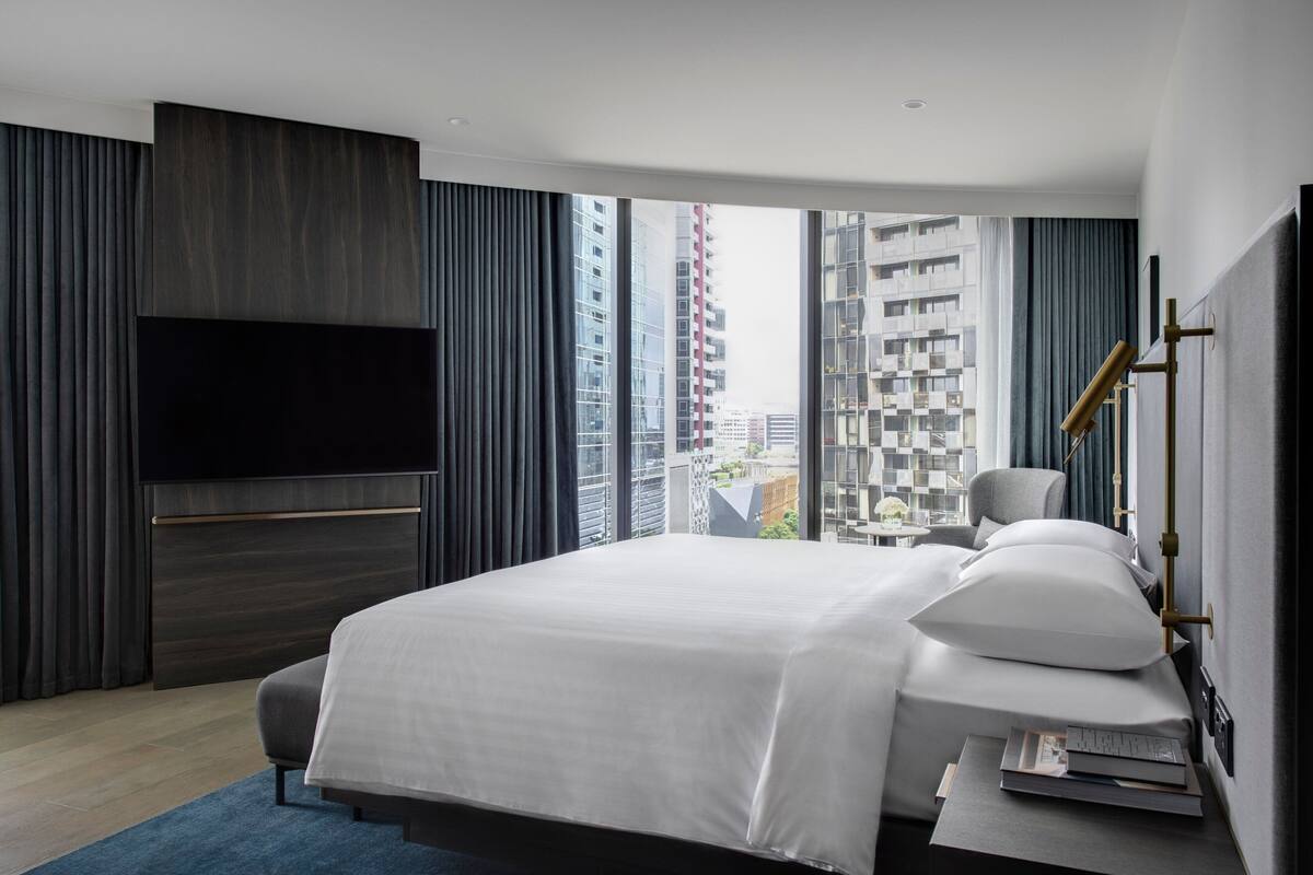 A modern hotel room with a large bed, a flat-screen TV mounted on the wall, and a view of the cityscape through large windows.