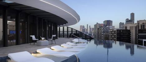 An infinity pool with white lounge chairs and a city skyline in the background.