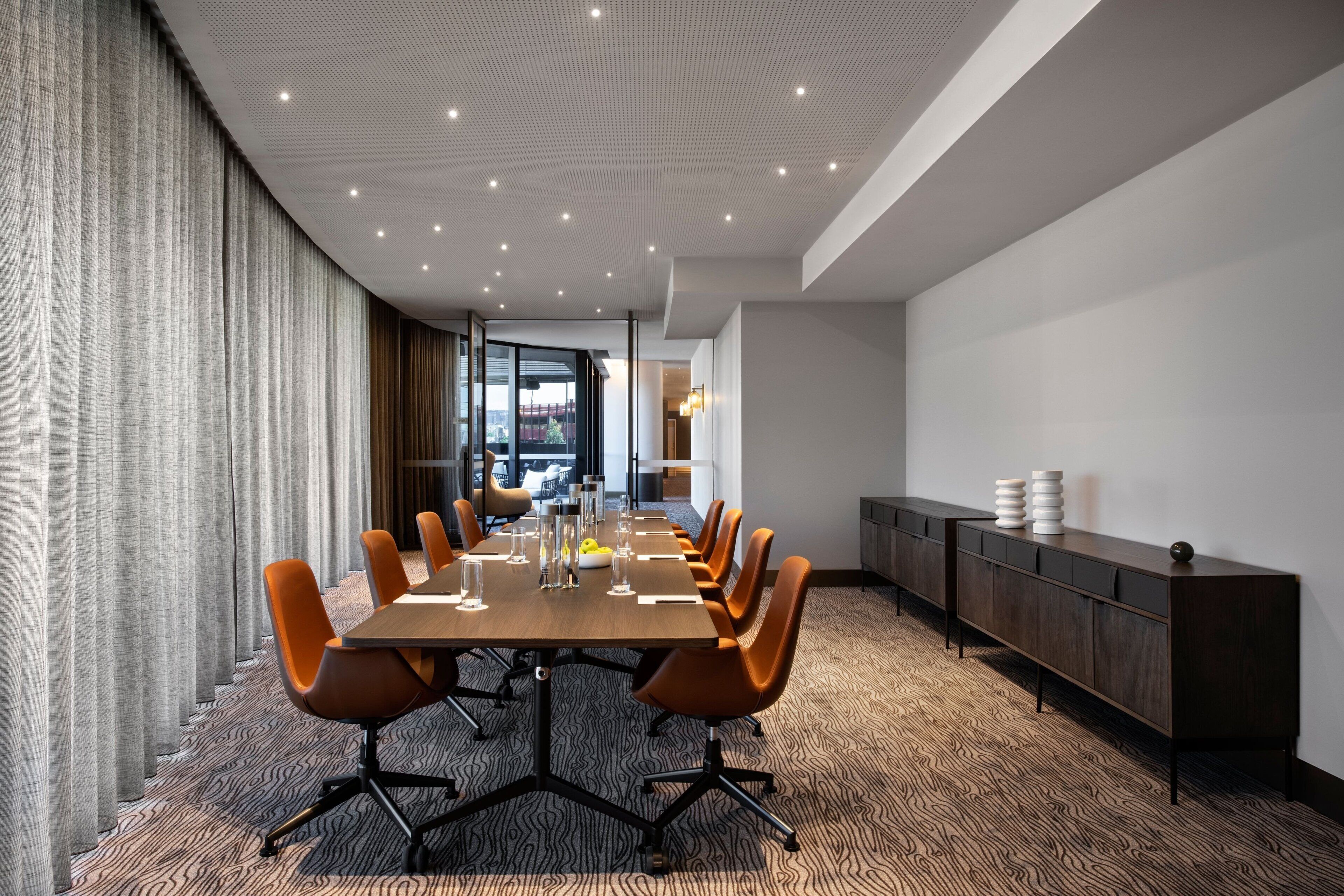 A conference room with a long table, orange chairs, a dark wooden cabinet, and patterned carpet.