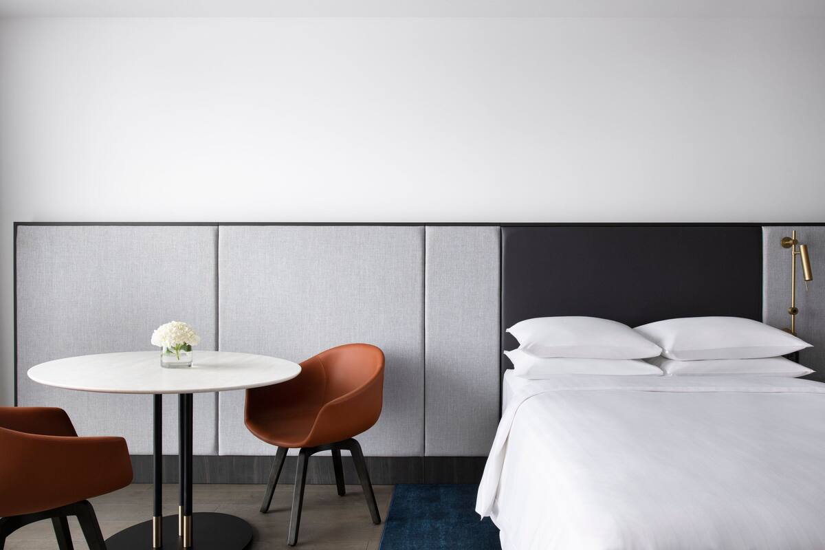 A modern hotel room with a bed, a round table with two chairs, and a small vase with flowers.