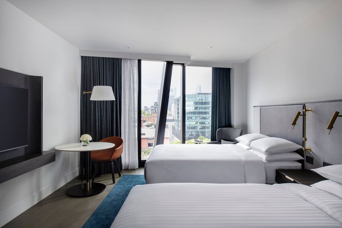 A modern hotel room with two beds, a desk, a chair, and a large window offering a city view.