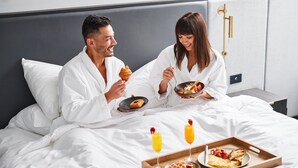 A couple in a hotel room with a bed, a tray with waffles, and a bowl of cereal.