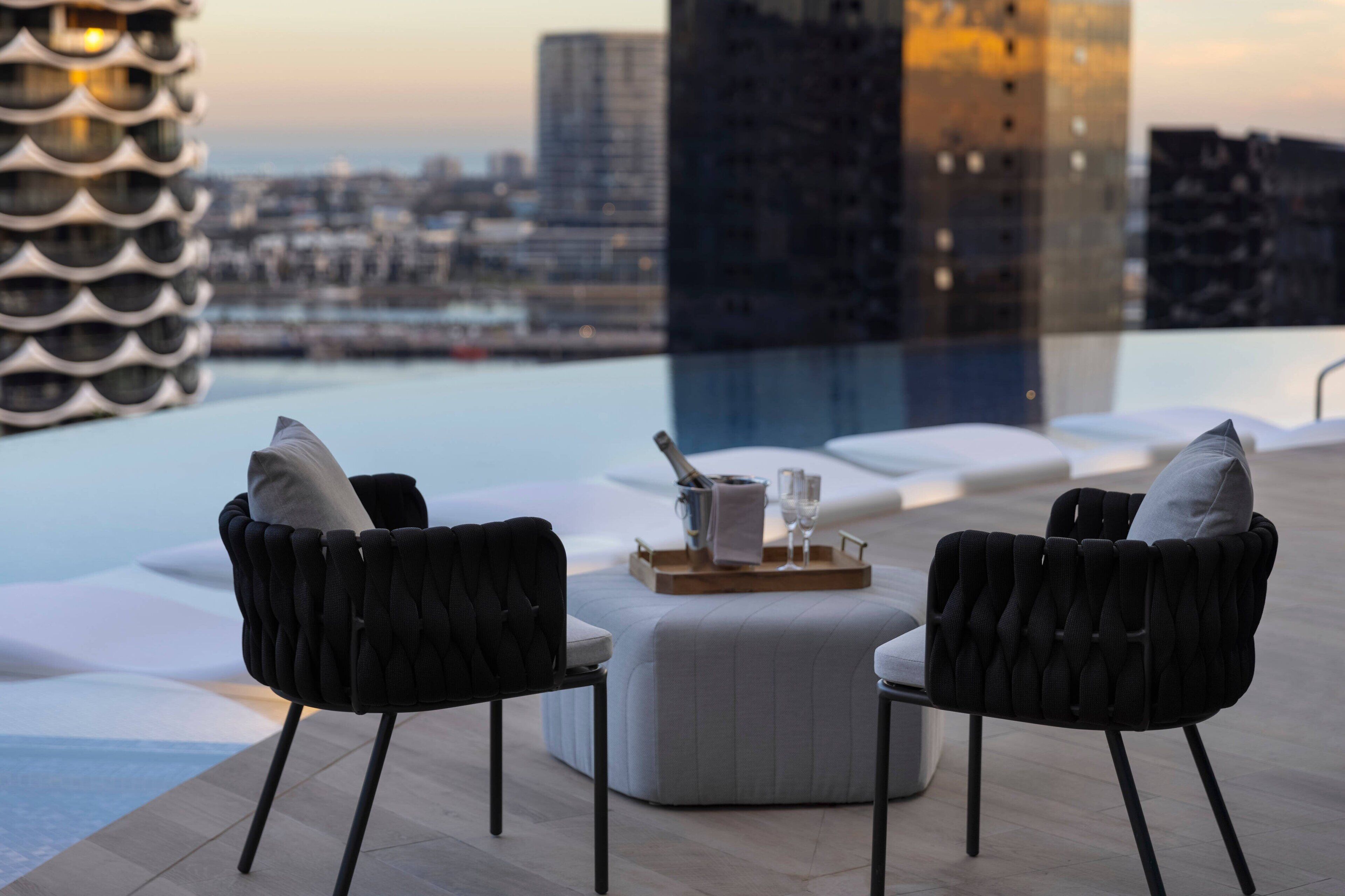 A rooftop terrace with two black upholstered chairs, a white ottoman, and a tray with a bottle and glasses. A modern building with a unique facade is visible in the background.