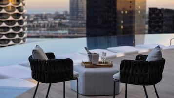 A rooftop terrace with two black upholstered chairs, a white ottoman, and a tray with a bottle and glasses. A modern building with a unique facade is visible in the background.